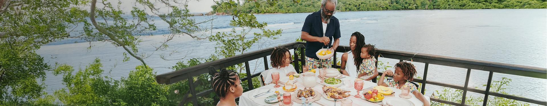 Family enjoying an outdoor meal on a deck overlooking water, with food served at a round table.