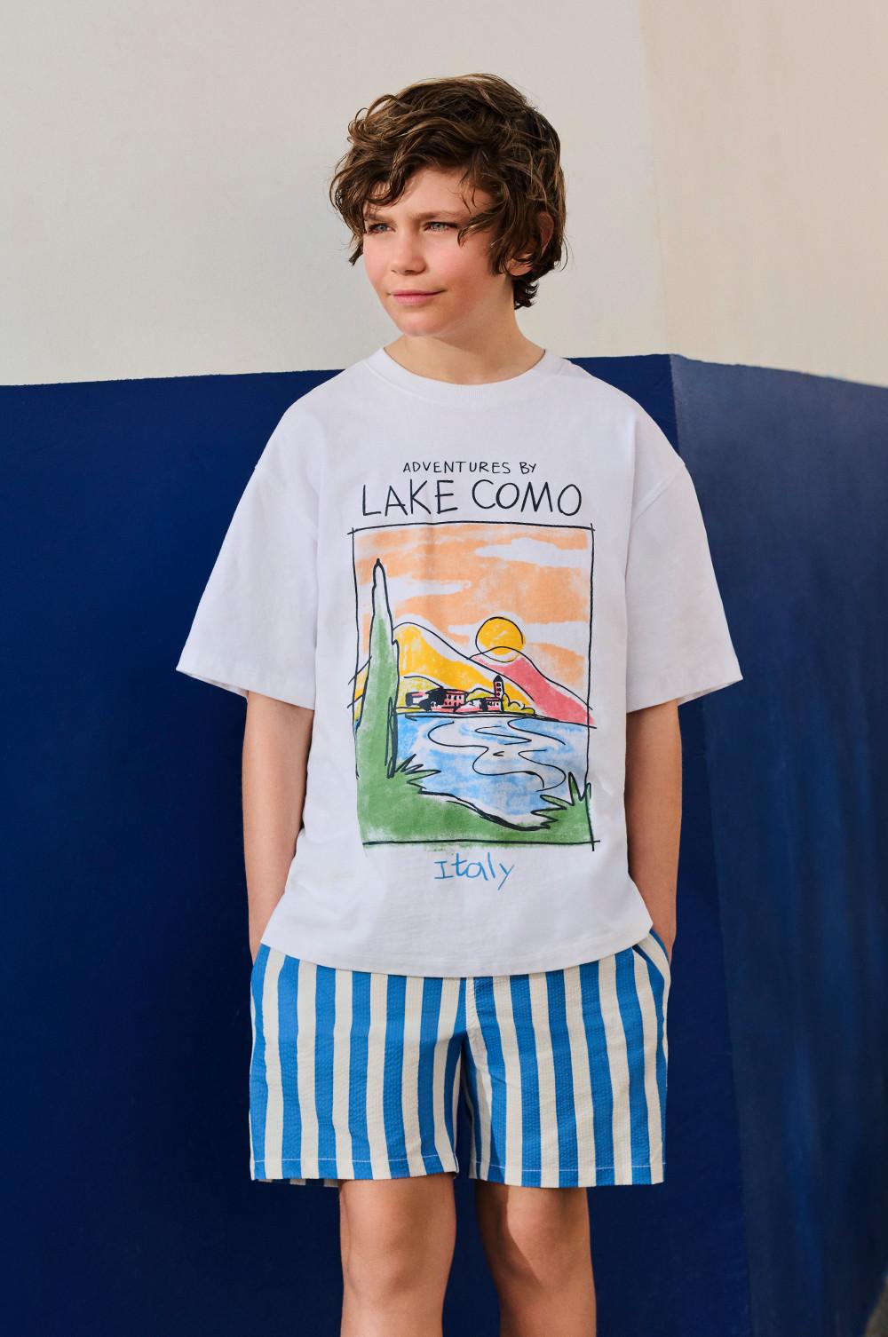 Boy wearing white t-shirt with LAKE COMO graphic print and blue striped shorts.