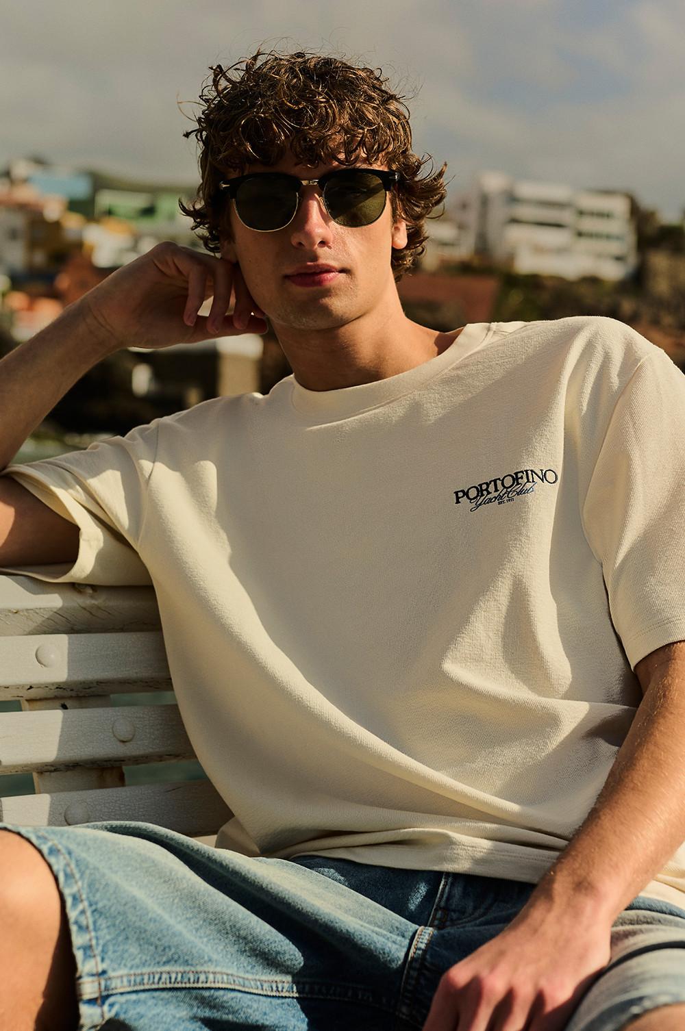 Model wearing cream PORTOFINO T-shirt and sunglasses, relaxing on a sun lounger outdoors.