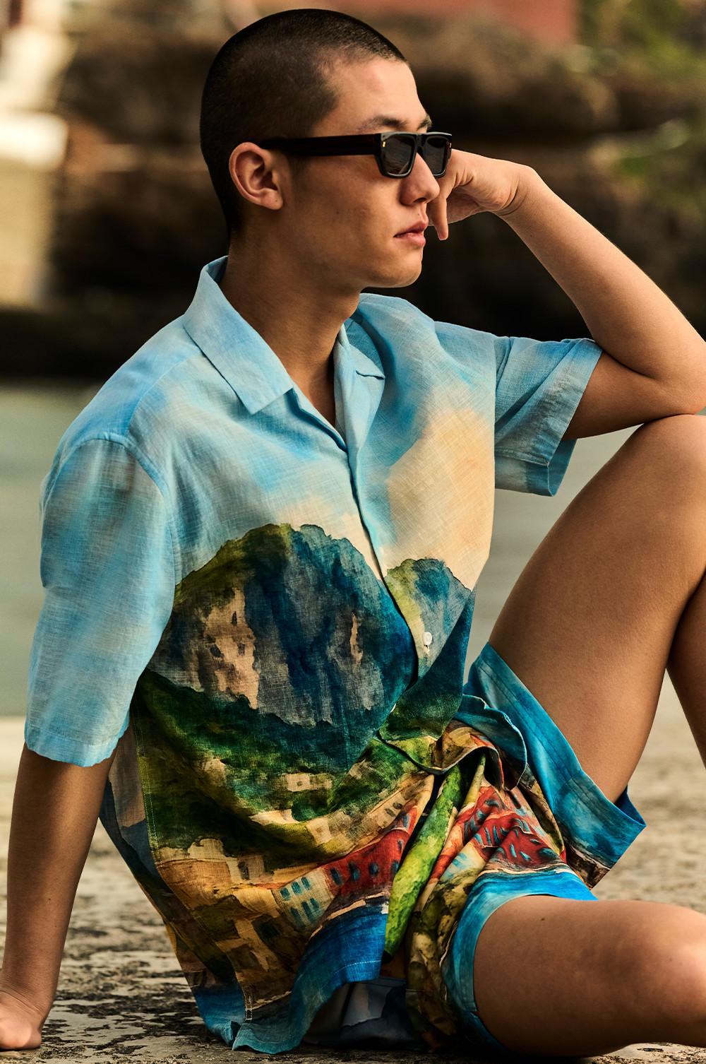 Model sitting on beach wearing black sunglasses and blue short-sleeved shirt with scenic landscape print.