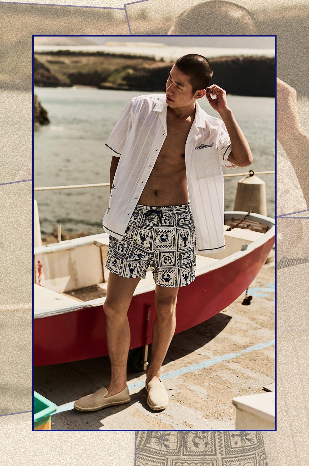Model wearing open white short-sleeved shirt and patterned swim shorts standing by boats at a coastal harbour.