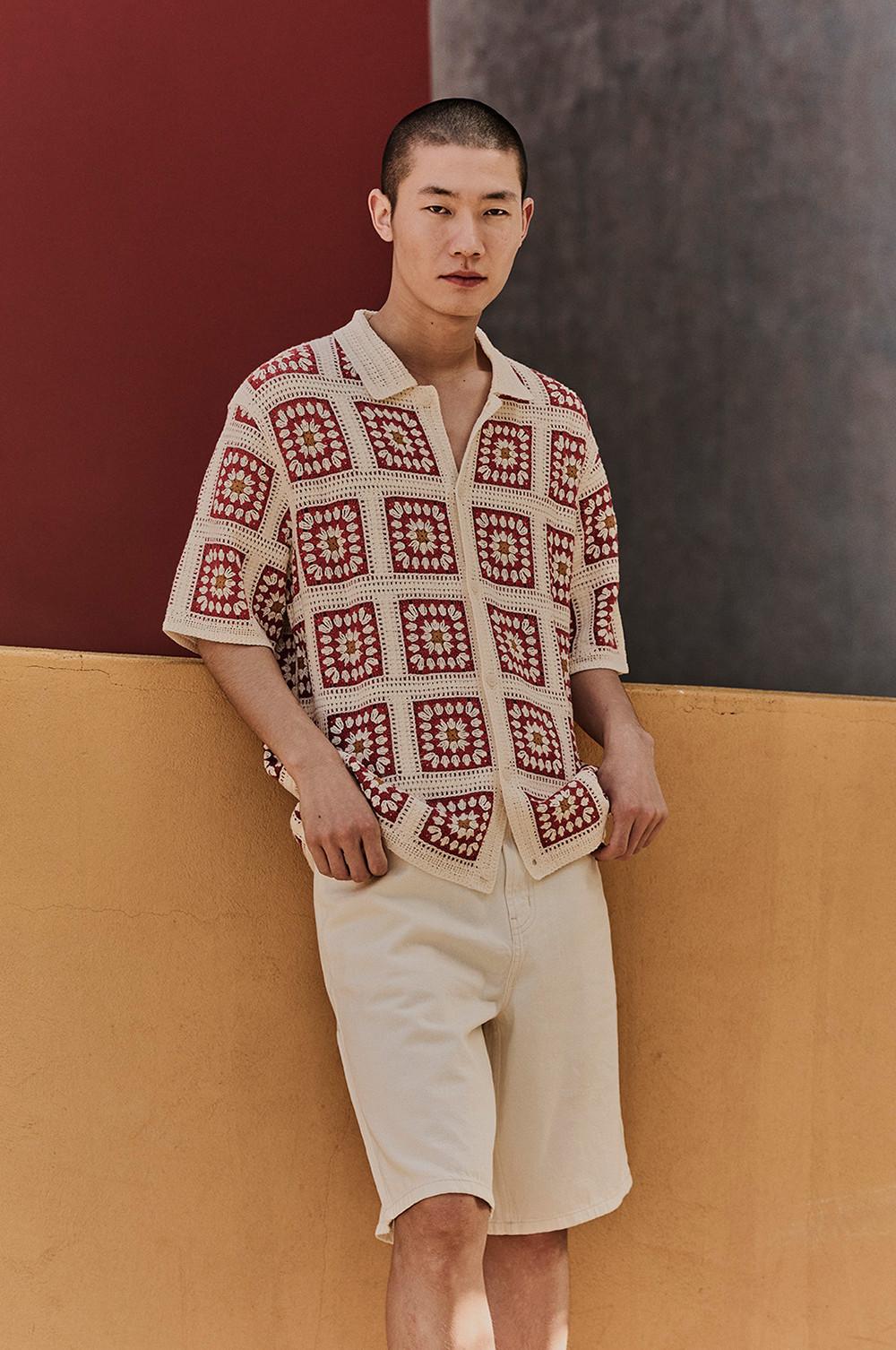 Model wearing crochet granny square shirt in burgundy and cream with beige shorts, leaning against a wall.