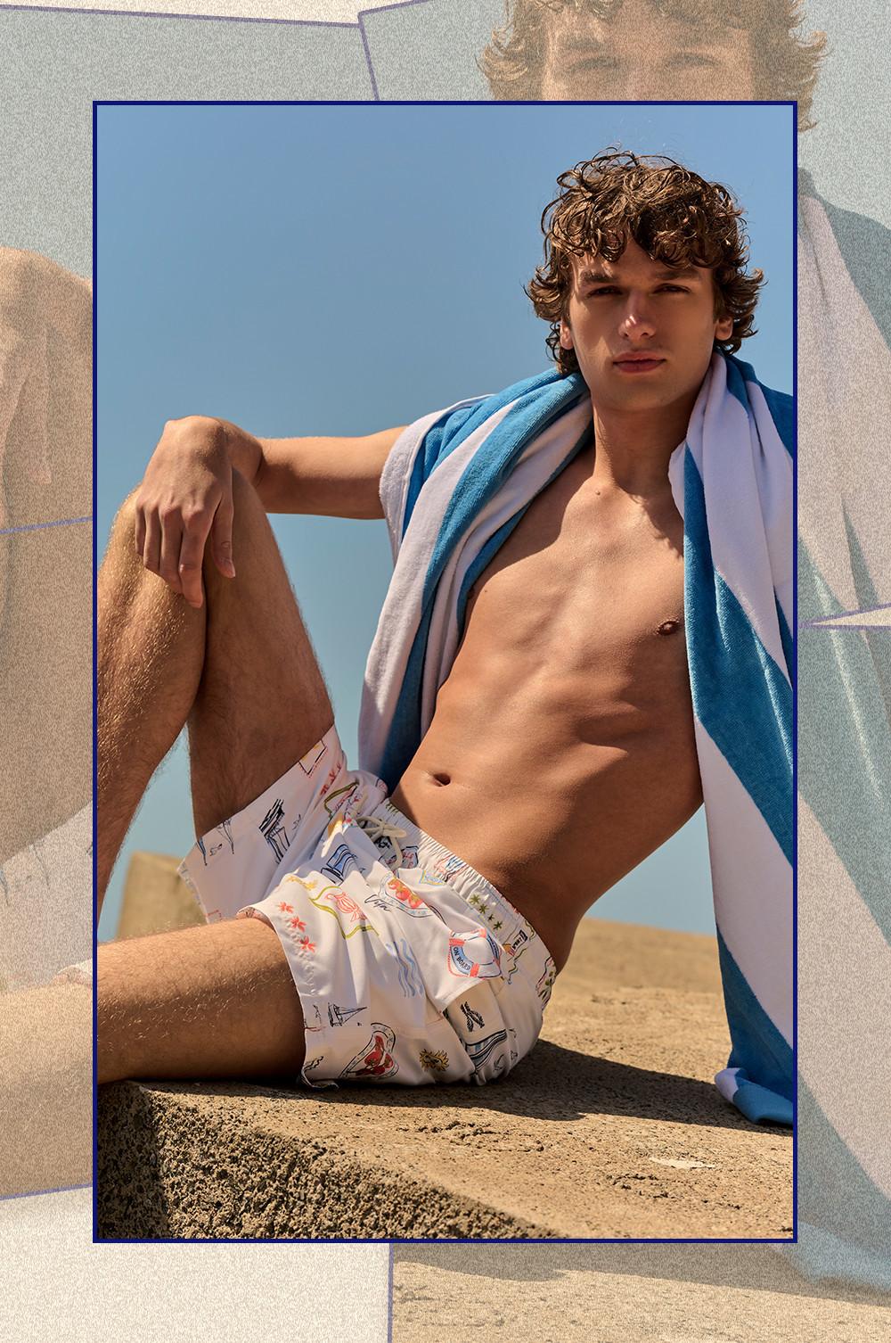 Model sitting outdoors wearing white printed swim shorts with a blue and white striped towel draped over shoulder.