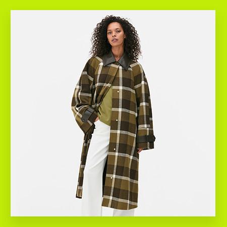 Person wearing oversized brown plaid coat with white trousers against white background.