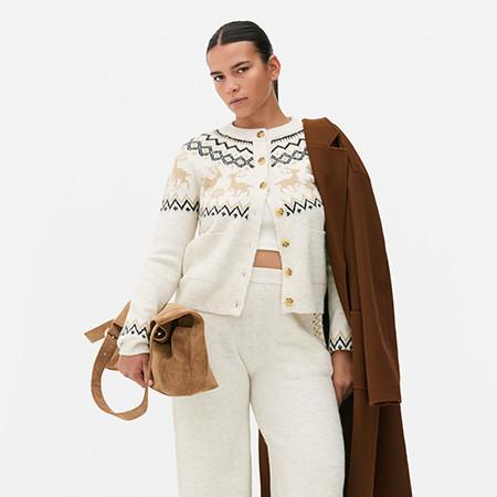 Person wearing cream fair isle cardigan with white trousers, holding brown coat and handbag.