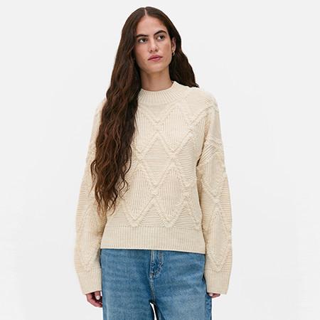 Person wearing cream cable-knit jumper with diamond pattern and blue jeans against white background.