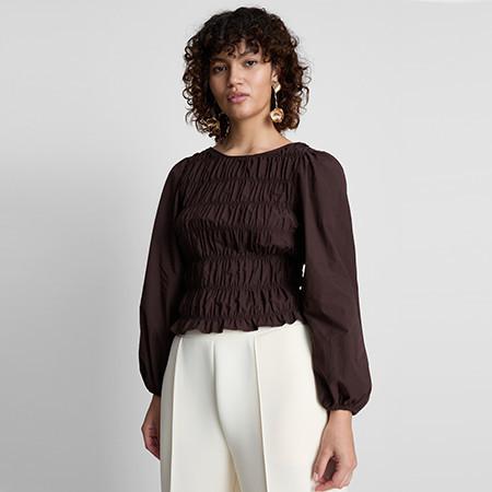 Brown ruched blouse with long balloon sleeves paired with white trousers against a grey background.