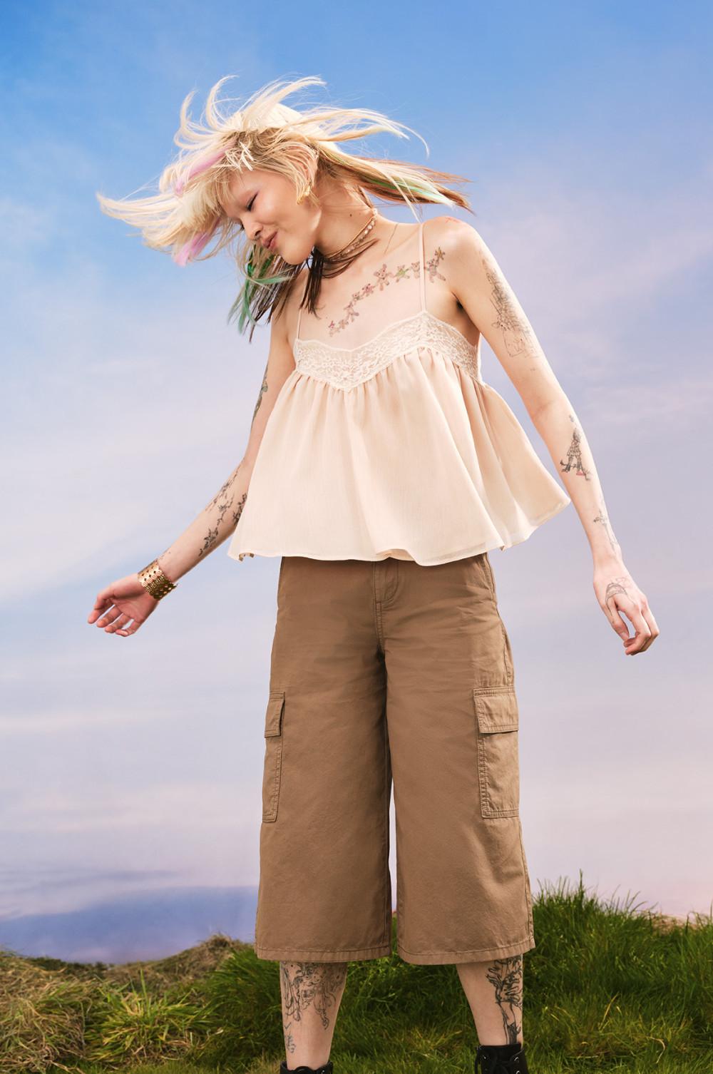 Woman with windswept hair wearing cream camisole top with lace detail and brown cargo trousers outdoors.