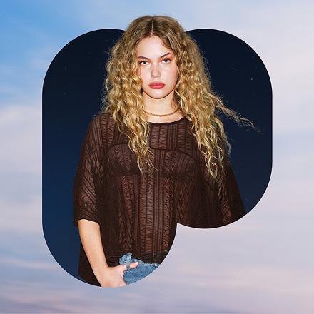 Woman with long blonde curly hair wearing a brown textured knit top with three-quarter length sleeves.