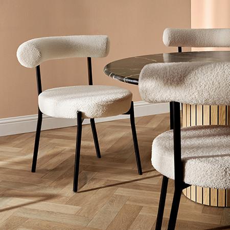 Modern dining chair with cream bouclé upholstery, curved backrest and black metal legs beside a marble table.