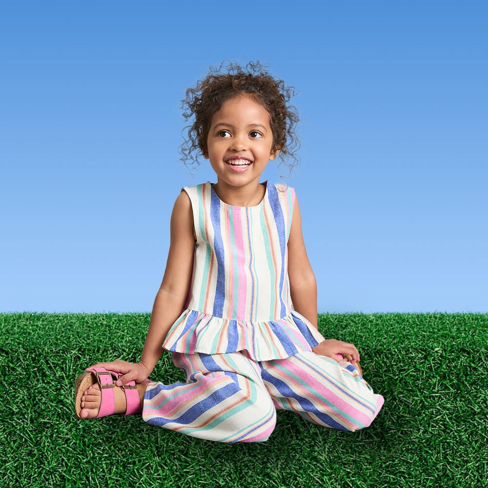 Young girl sitting on grass wearing a multicolored striped sleeveless dress with ruffle hem detail.
