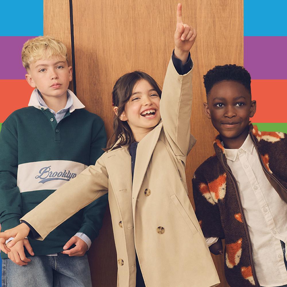Three children wearing casual outerwear including a Brooklyn rugby shirt, beige trench coat, and patterned sleeveless jacket.