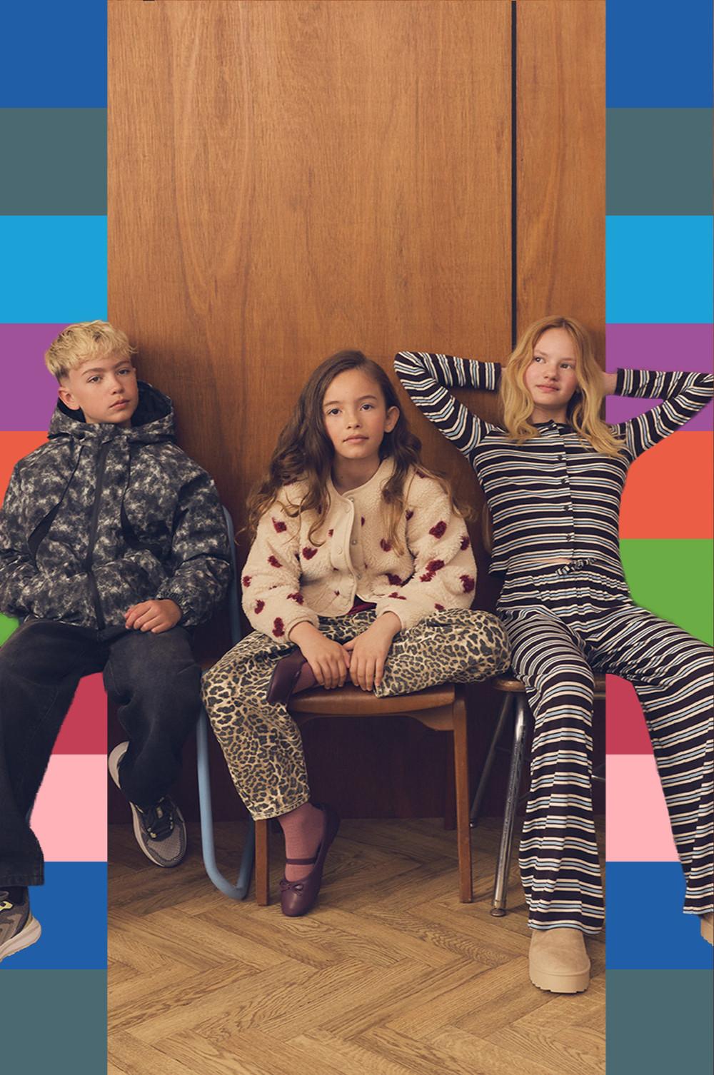 Three children sitting together wearing patterned clothing including a camouflage jacket, floral sweater and striped outfit.