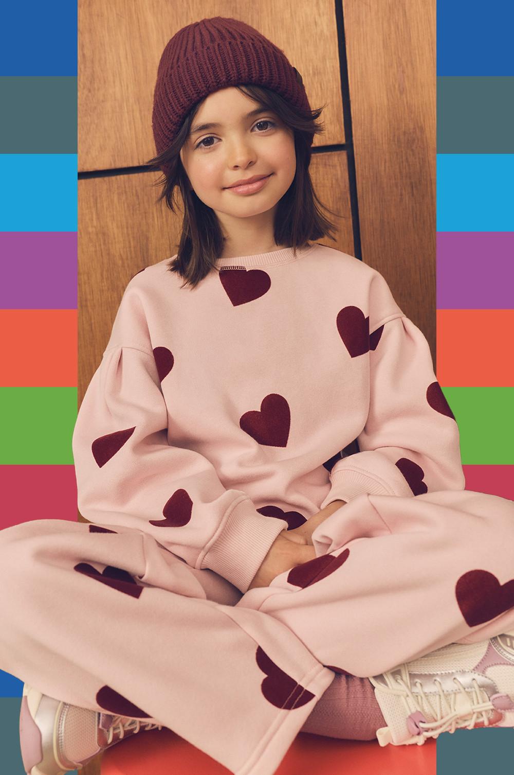 Child wearing pink heart-print sweatshirt and matching joggers with burgundy knitted beanie.