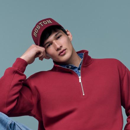 Man wearing burgundy quarter-zip sweatshirt and matching Boston cap with hand raised to head.