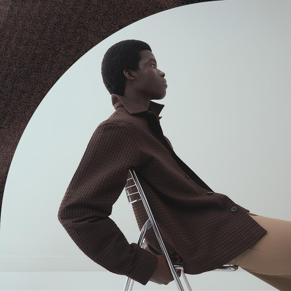 Model seated on chair wearing brown textured jacket with high collar in profile view.