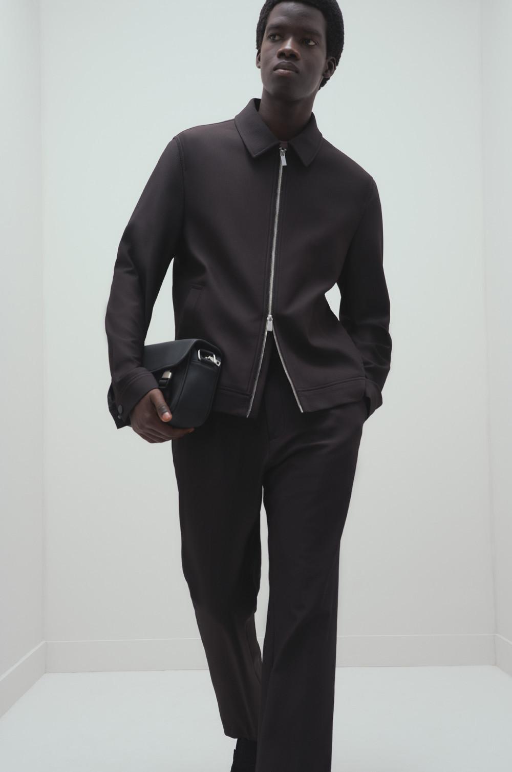 Model wearing black collared jacket with full-length zip fastening and matching pants, holding a helmet.