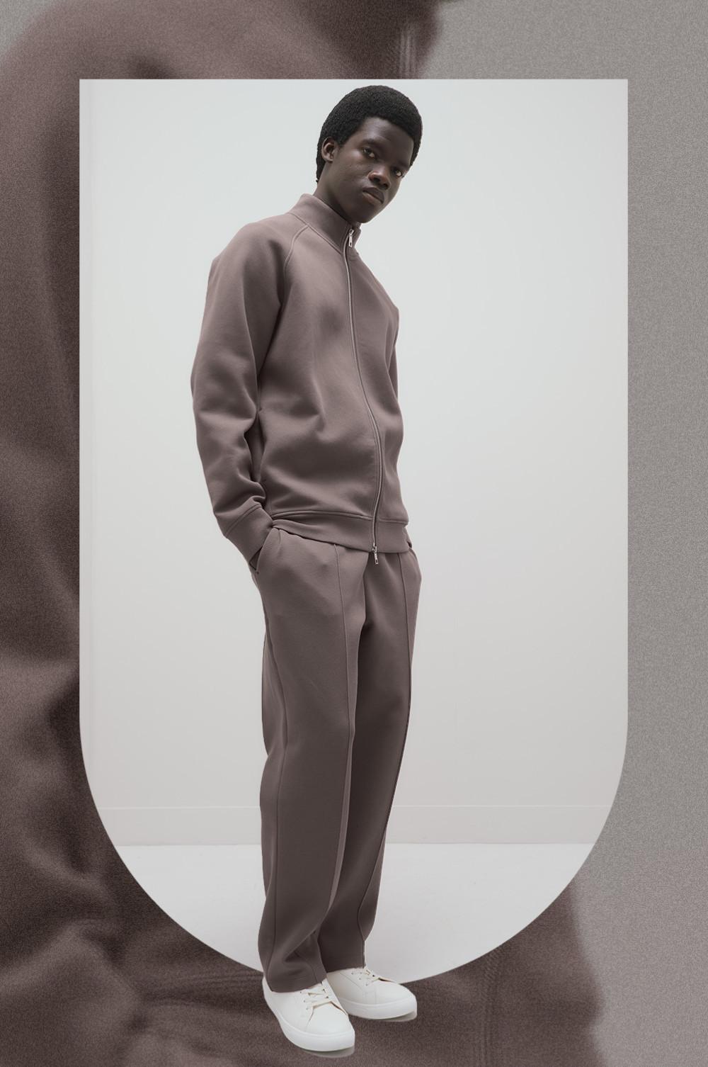 Man wearing taupe zip-up tracksuit with matching pants and white sneakers, paired with a black beanie.