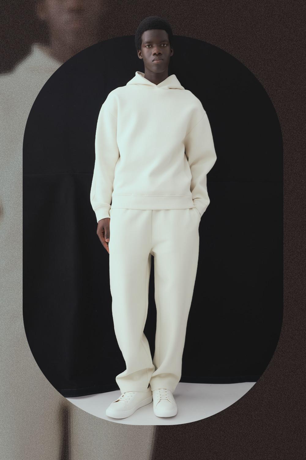 Model wearing cream hoodie and matching joggers with white sneakers, standing against dark archway.