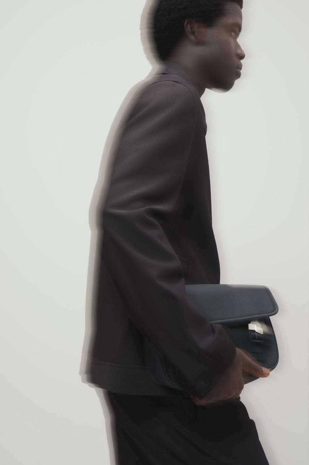 Model in profile wearing dark high-neck top, carrying a black structured handbag under their arm.