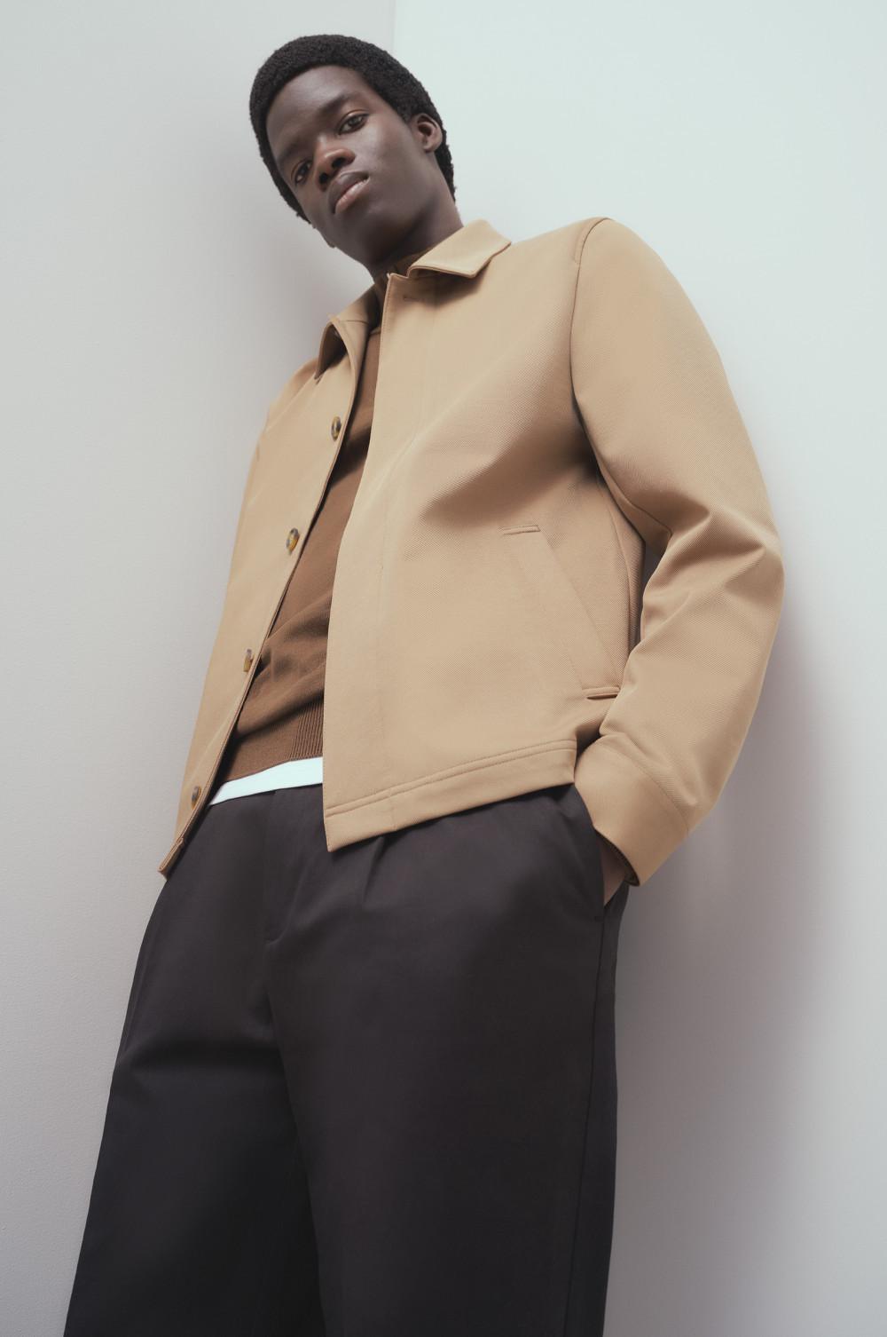 Model wearing a camel-colored jacket with button fastening, brown sweater and black pants.