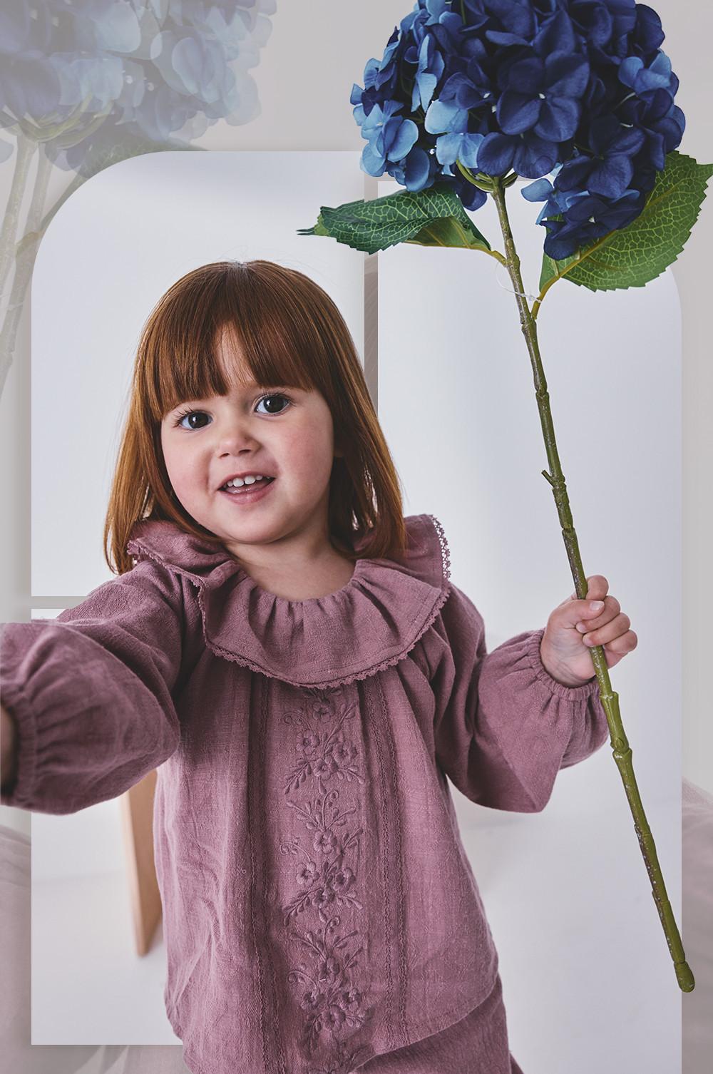 Young girl smiling in a mauve embroidered dress with ruffled collar, holding a blue hydrangea flower.
