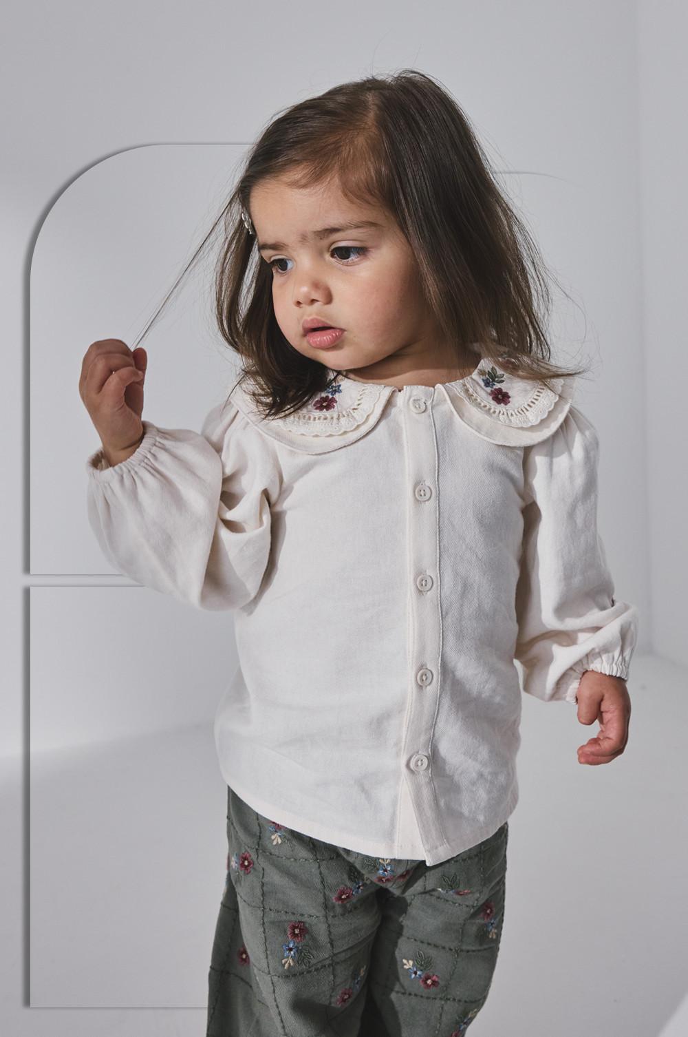 Young girl wearing white blouse with embroidered collar and floral pants.