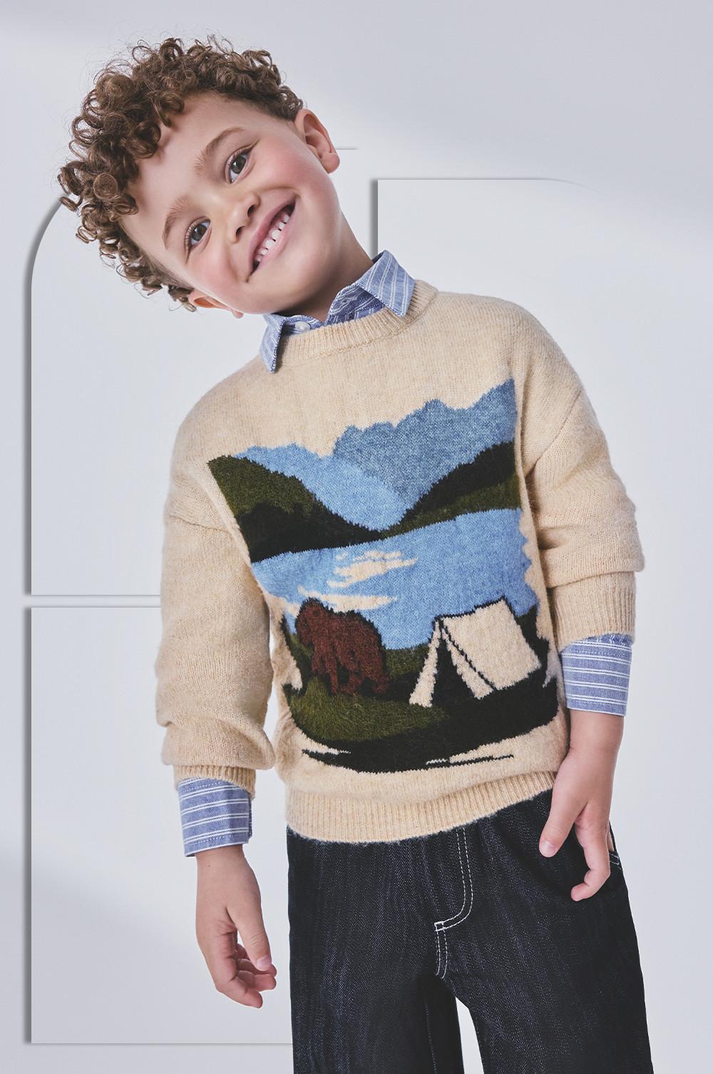Child smiling and tilting head, wearing beige sweater with landscape intarsia design over striped shirt.