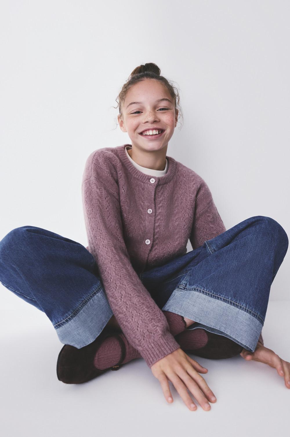 Young girl smiling whilst sitting cross-legged, wearing a mauve knitted cardigan and blue denim jeans.