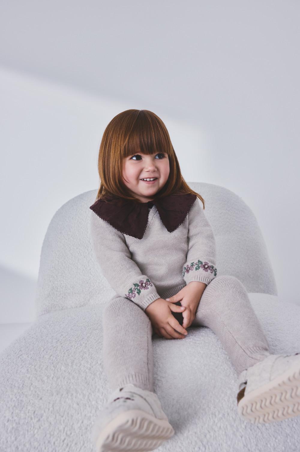 Young child smiling whilst sitting in a white chair, wearing a gray knitted sweater with brown collar.