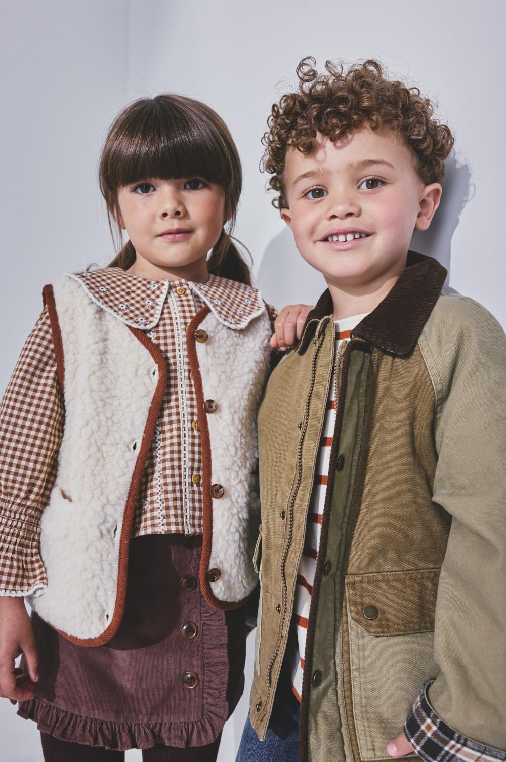Two children smiling together, wearing fall layered outfits with vests and gingham details.