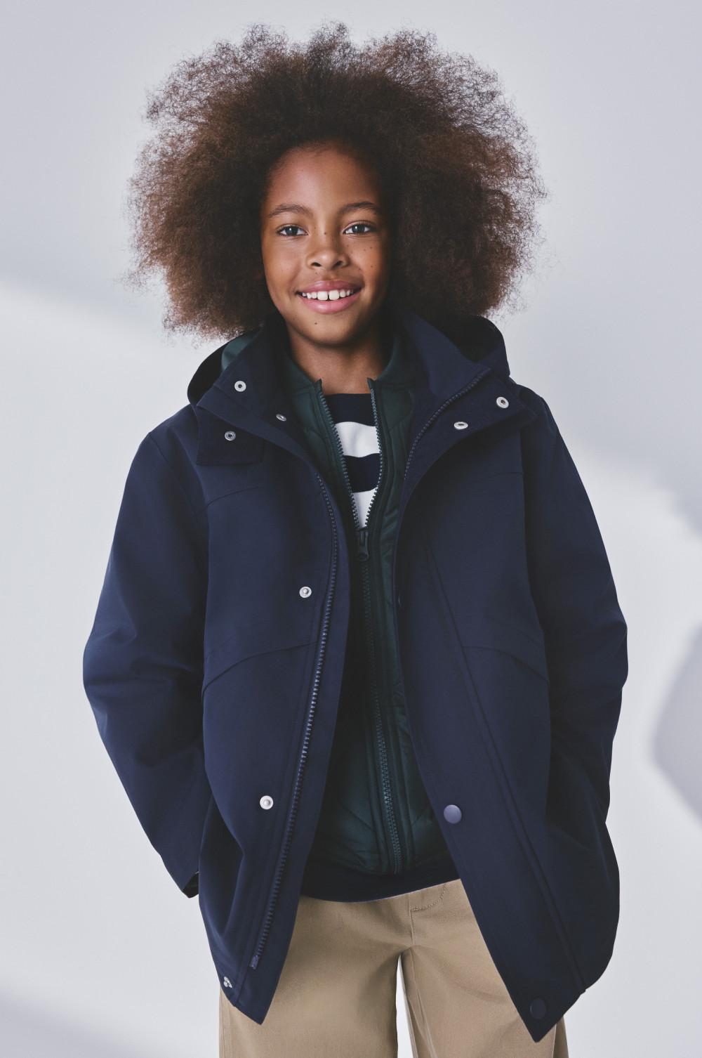 Child wearing navy hooded jacket with zip fastening and beige pants, smiling at camera.