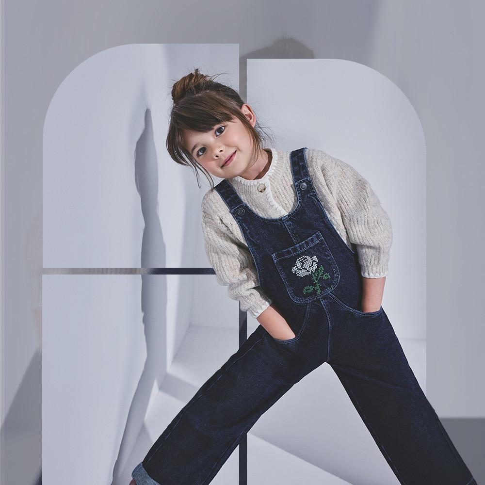 Child wearing denim dungarees with embroidered pocket detail and cream knitted sweater, posing playfully.