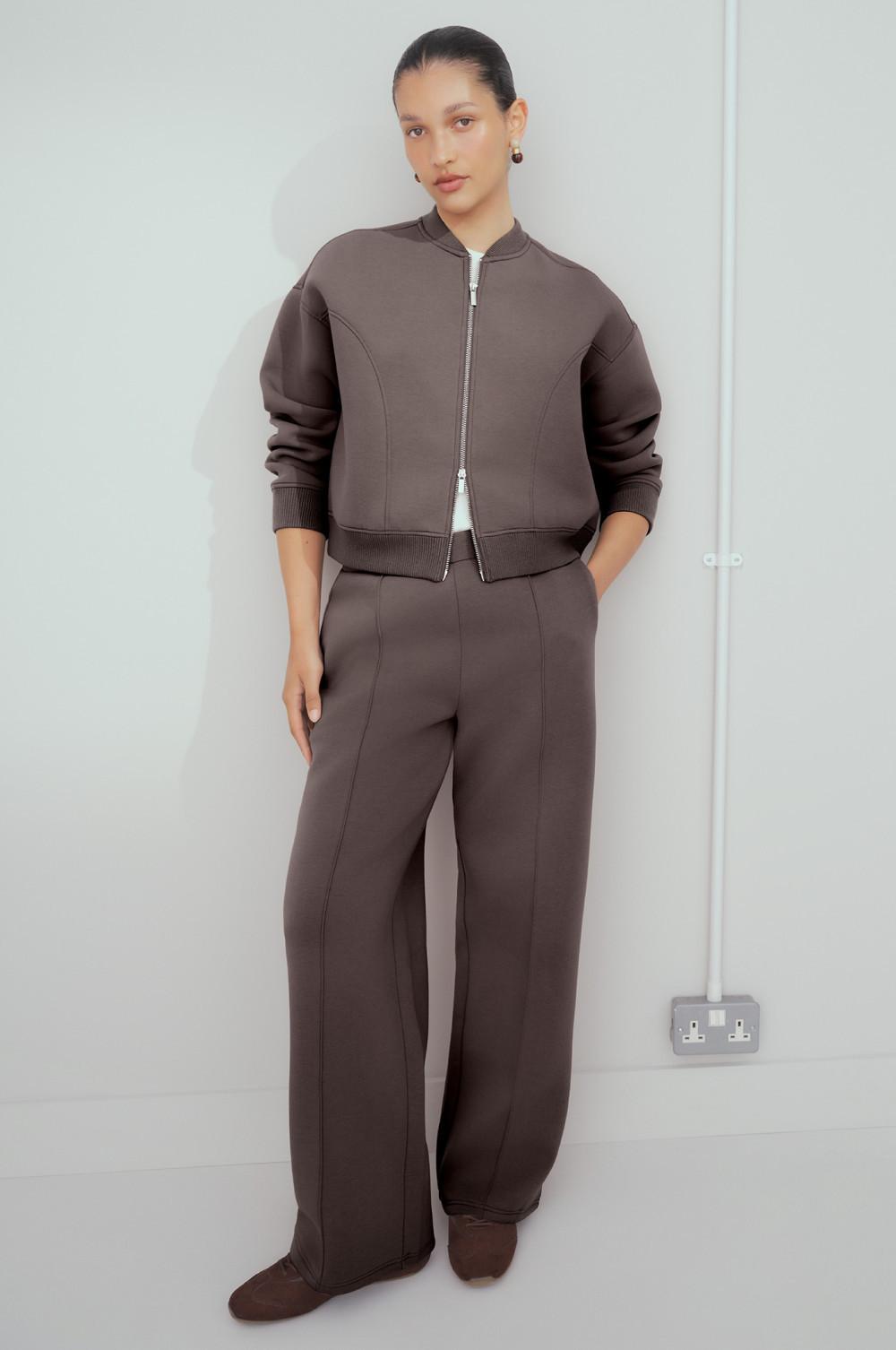 Model wearing a brown zip-up bomber jacket and matching wide-leg pants in a coordinated tracksuit.