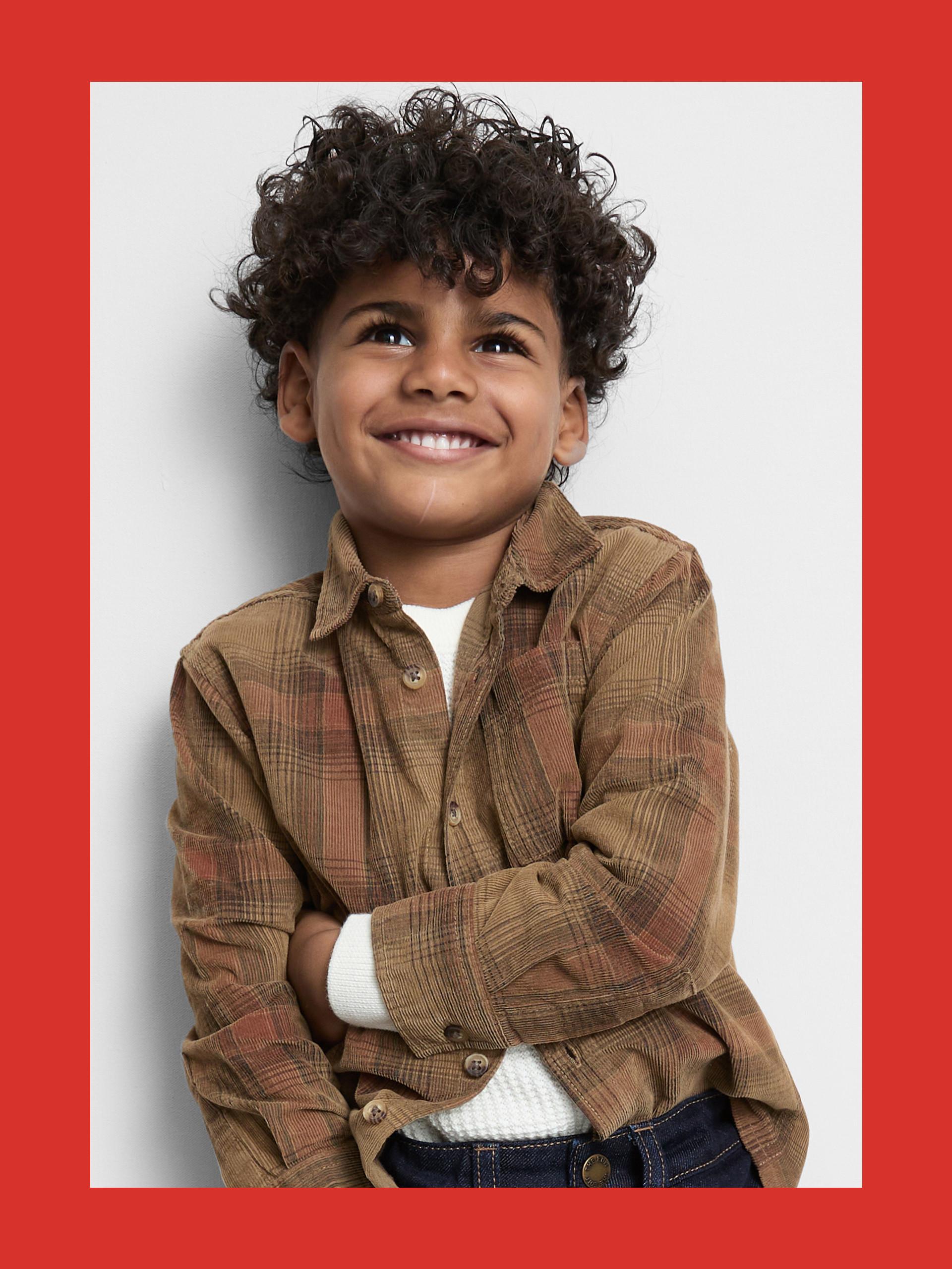 Child wearing brown corduroy plaid shirt over white top with curly dark hair, smiling and looking upward.