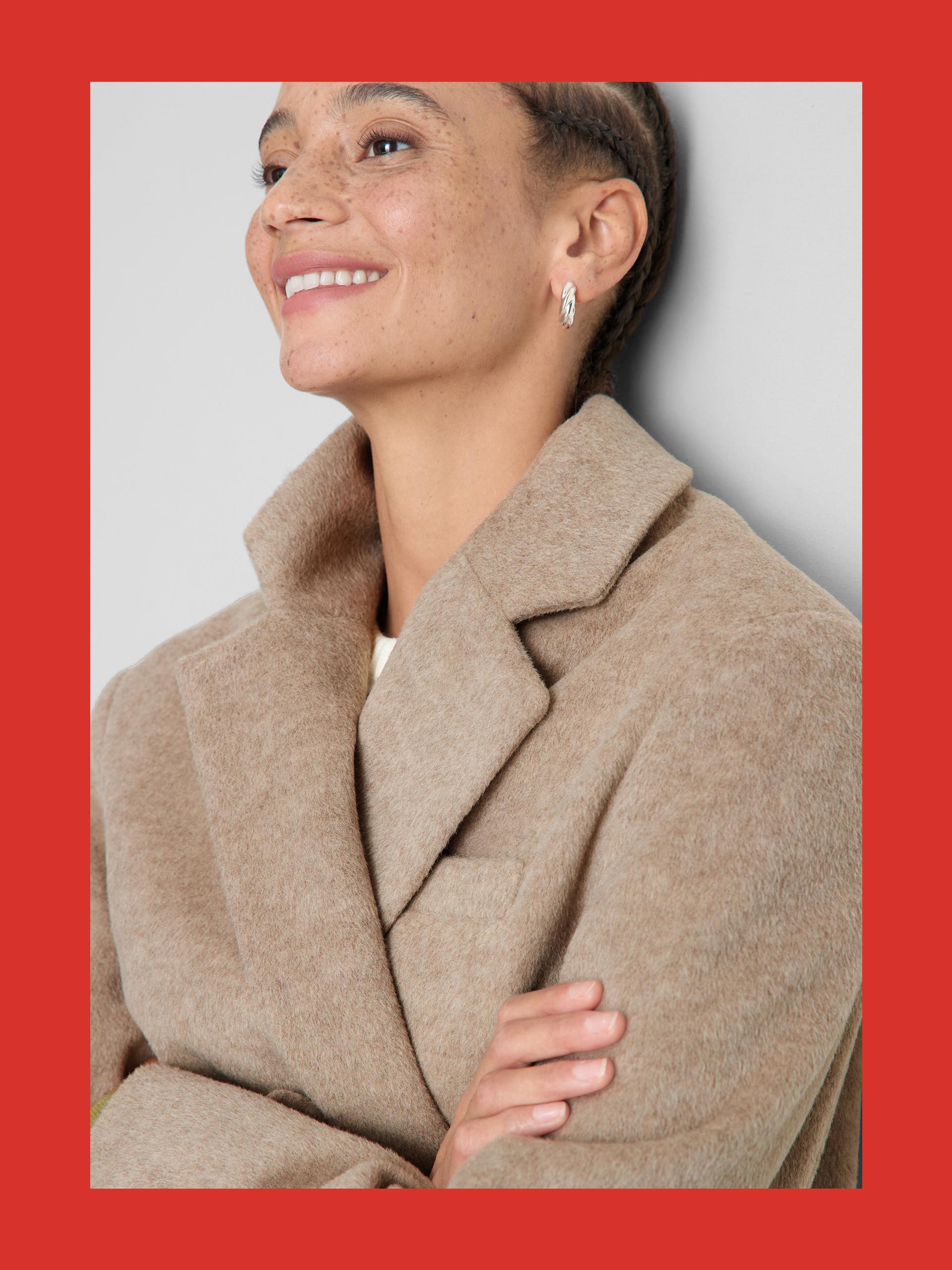 Person wearing a beige woollen coat with lapels and small silver hoop earrings, smiling against a red background.
