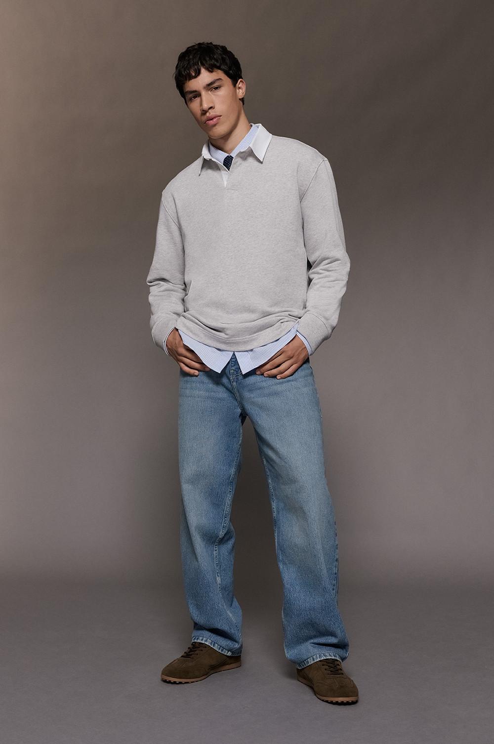 model wearing light gray crew neck sweater over striped shirt and tie with blue jeans and brown shoes.