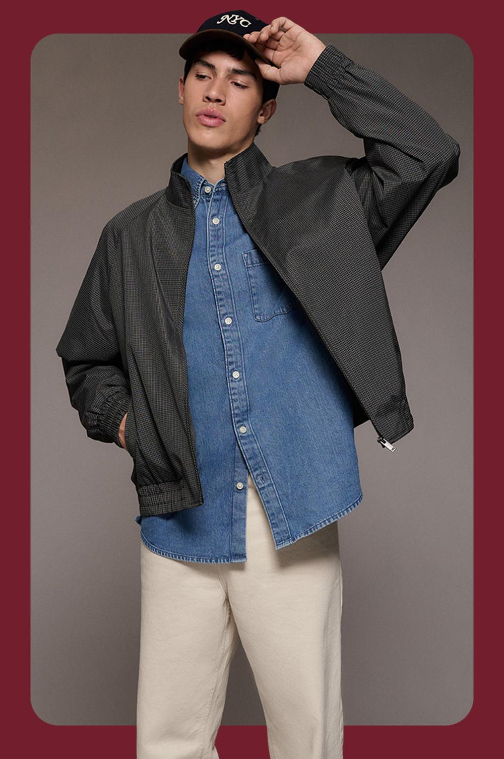 model wearing dark gray textured bomber jacket over denim shirt, beige pants and black cap, hand to head.