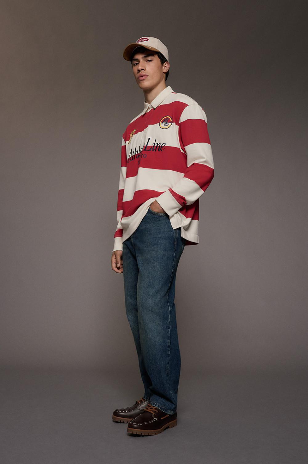 model wearing red and cream striped rugby shirt with blue jeans, baseball cap and brown boots.