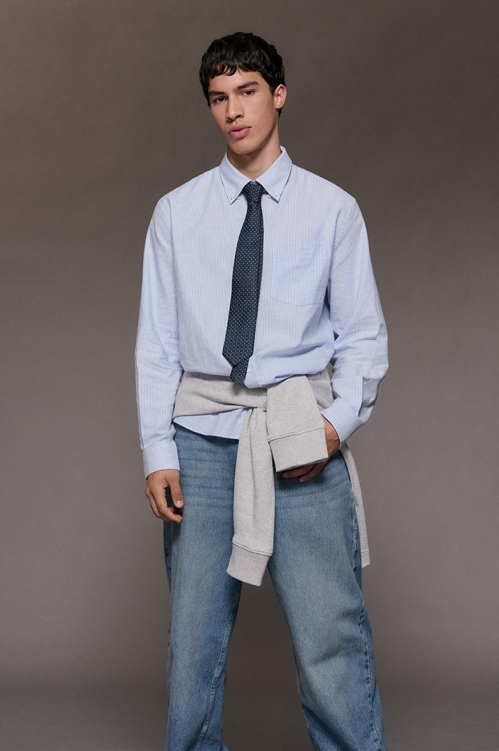 model wearing light blue striped shirt with navy polka dot tie, beige sweater tied at waist and blue jeans.