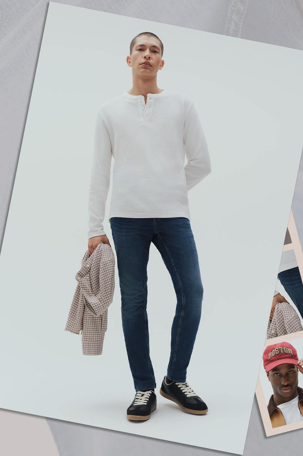 Model wearing white textured long-sleeve top with dark jeans and black sneakers, holding a checked shirt.