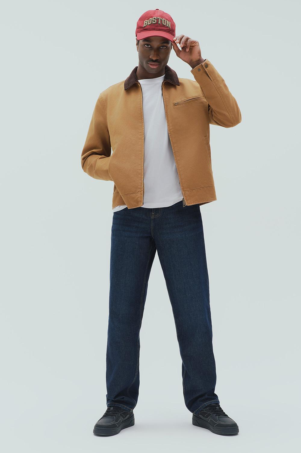 Model wearing tan jacket with brown collar, white T-shirt, dark jeans and red Boston cap, adjusting cap.