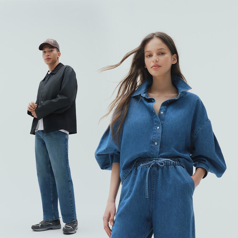 Woman in blue denim shirt and pants, man in black jacket and jeans with cap standing behind.