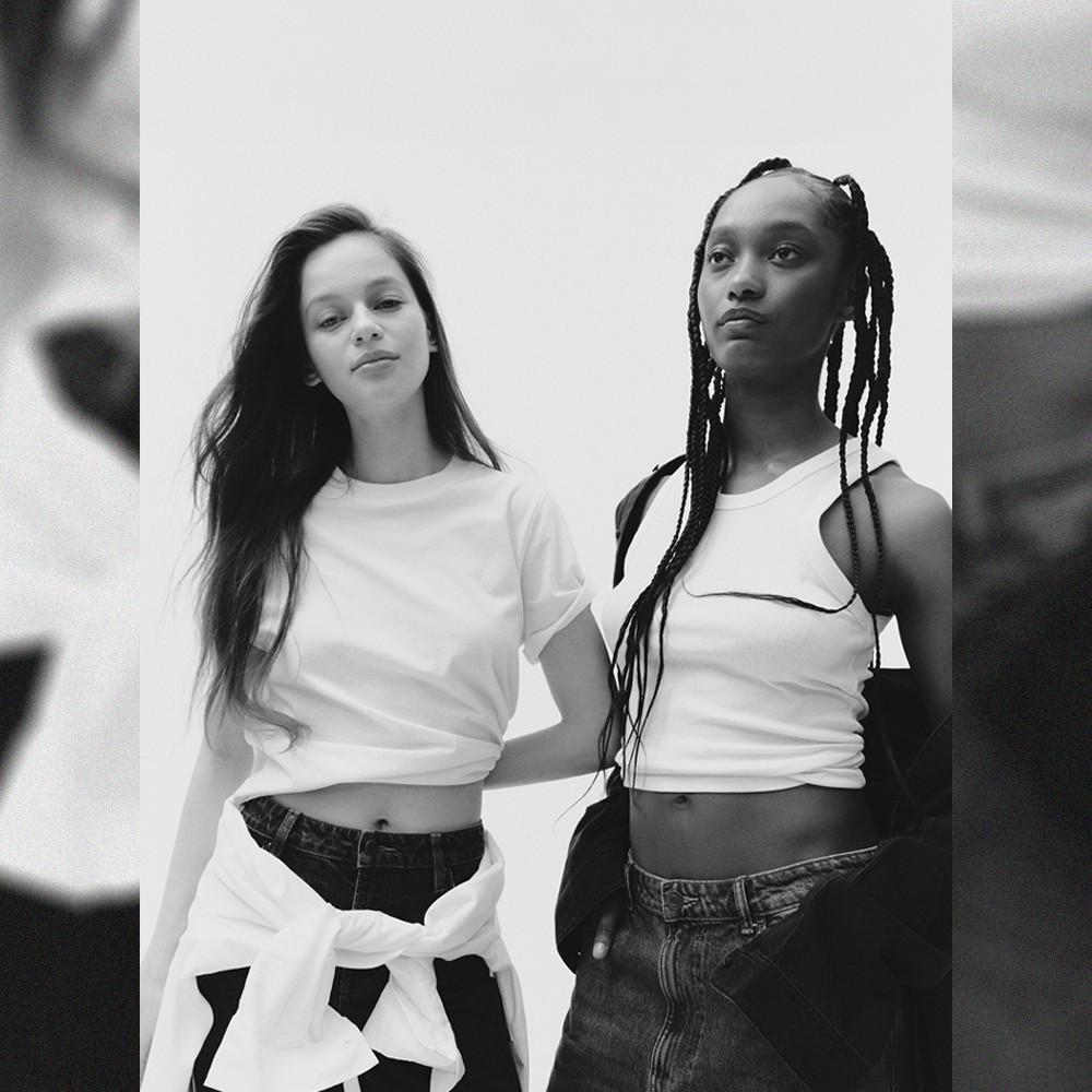 Two models standing side by side wearing white cropped T-shirts and jeans in a black and white photograph.