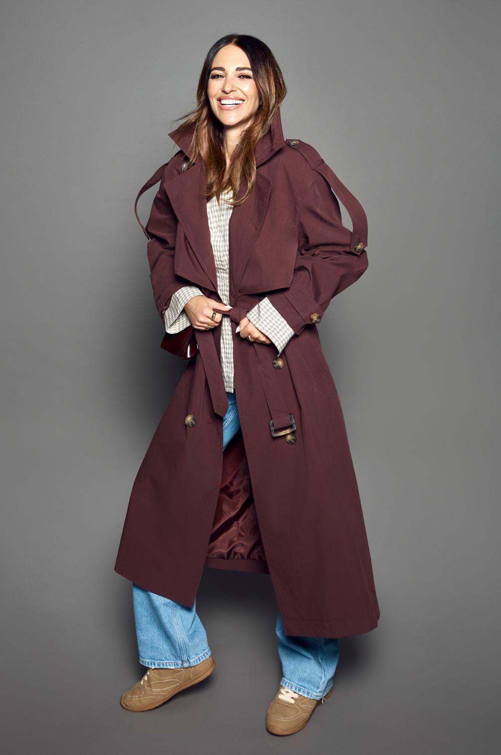 Model wearing burgundy trench coat over checked shirt and blue jeans with tan sneakers, smiling.