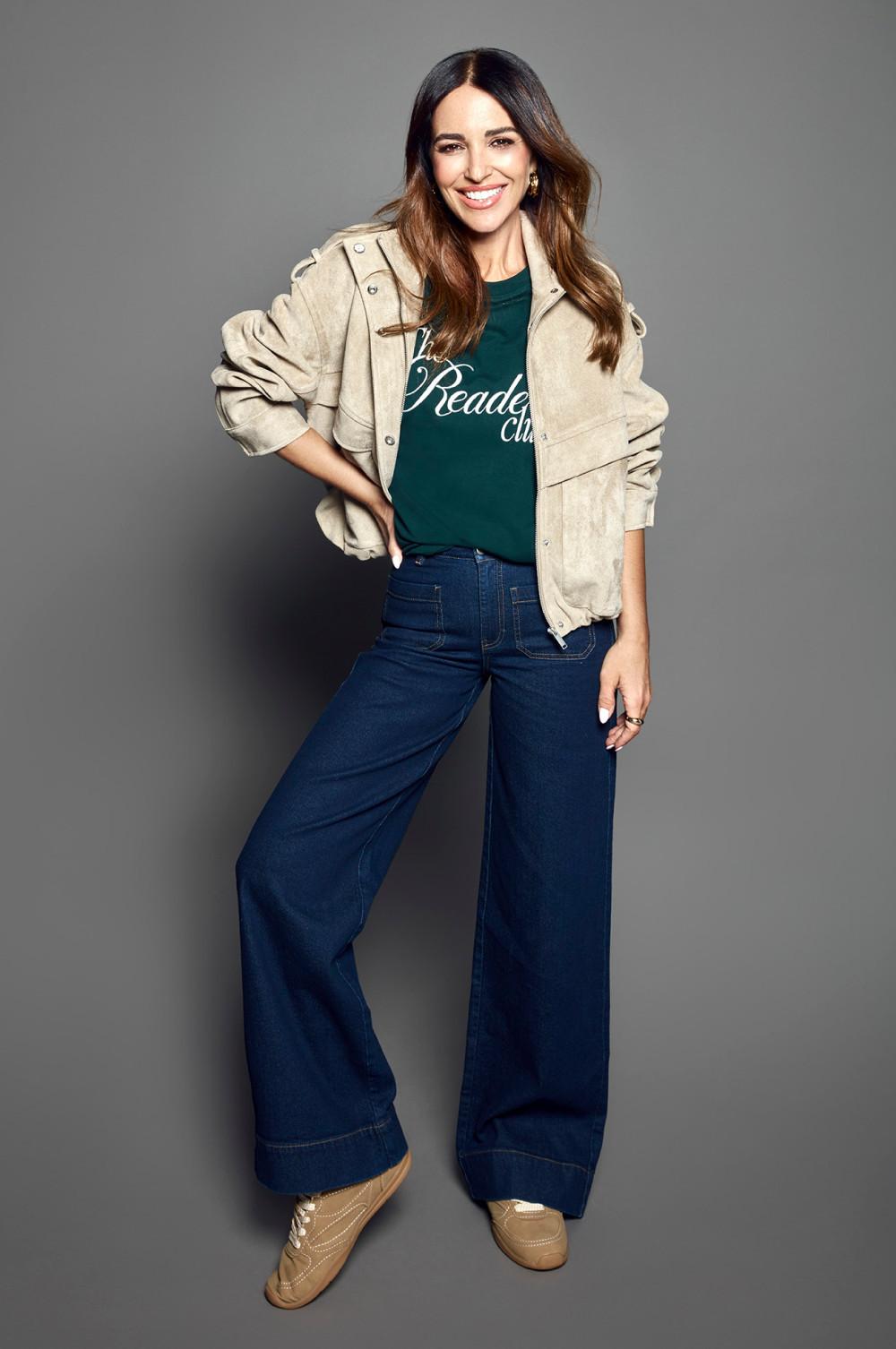 Woman smiling wearing beige jacket, green The Readers Club T-shirt, and dark wide-leg jeans with sneakers.