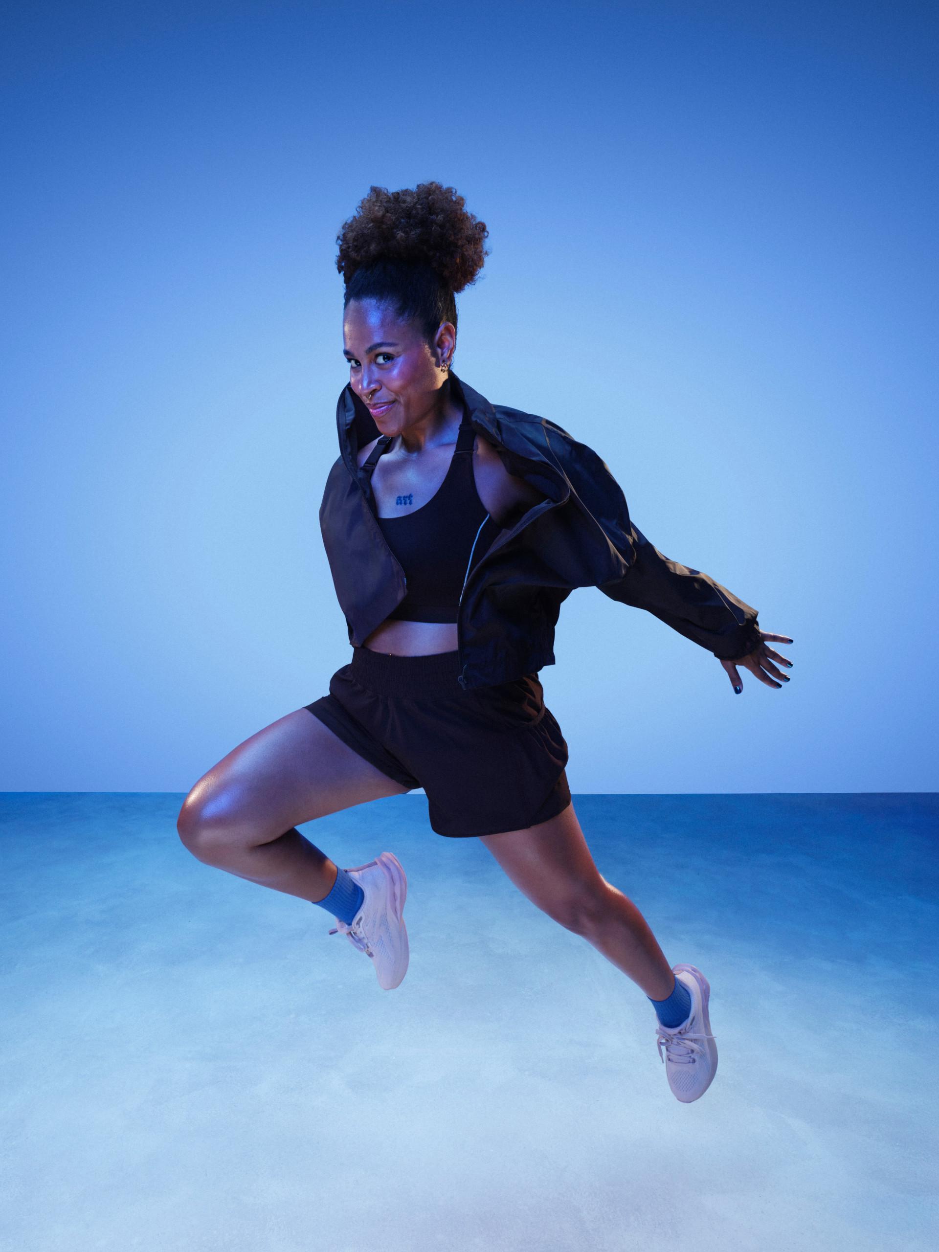 Modelo saltando enérgicamente con un conjunto de ropa deportiva negra que incluye sujetador deportivo, pantalón corto, chaqueta y zapatillas deportivas blancas.