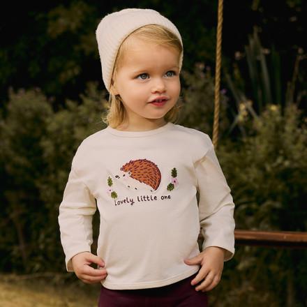 Young child wearing white long-sleeved top with pink and red heart print, sitting beside pink cushion.