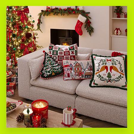Cosy living room decorated for Christmas with festive cushions, lit tree, stocking on fireplace and scented candles.