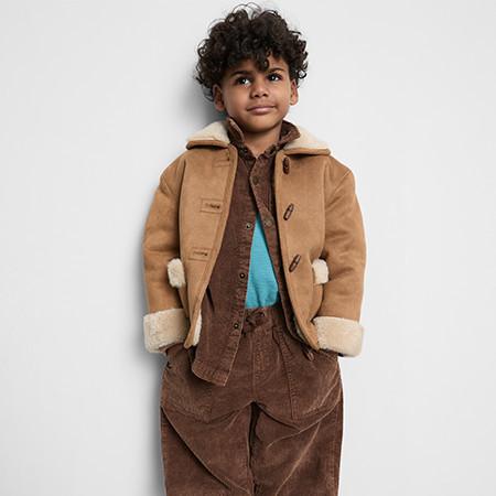 Child wearing tan duffle coat with shearling trim over brown corduroy outfit and blue top.